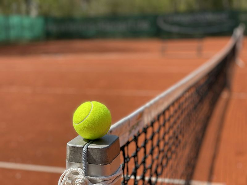 Gul tennisball balanserer på nettstolpen ved en rød grusbane