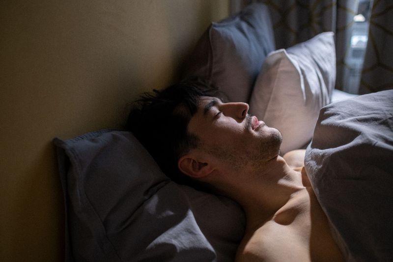 Mann sover på ryggen med munnen lett åpen i et svakt opplyst soverom