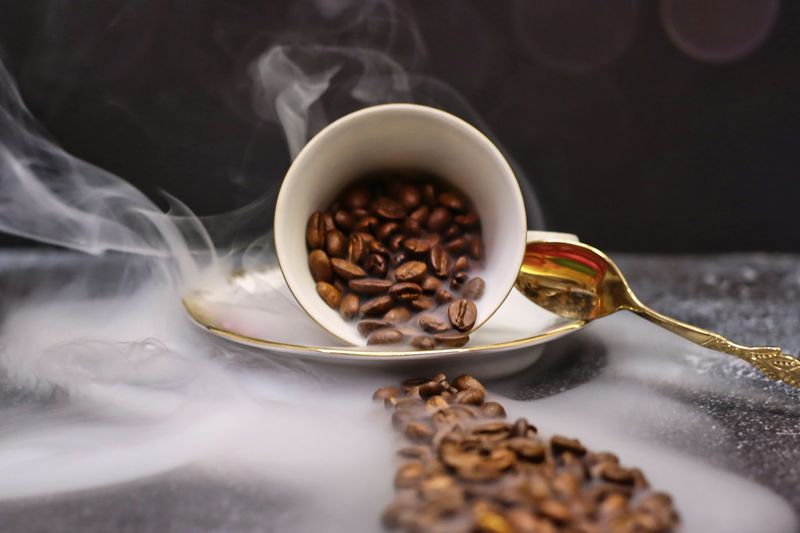 Kaffebønner velter ut av en hvit kopp med stigende damp og en gyllen skje