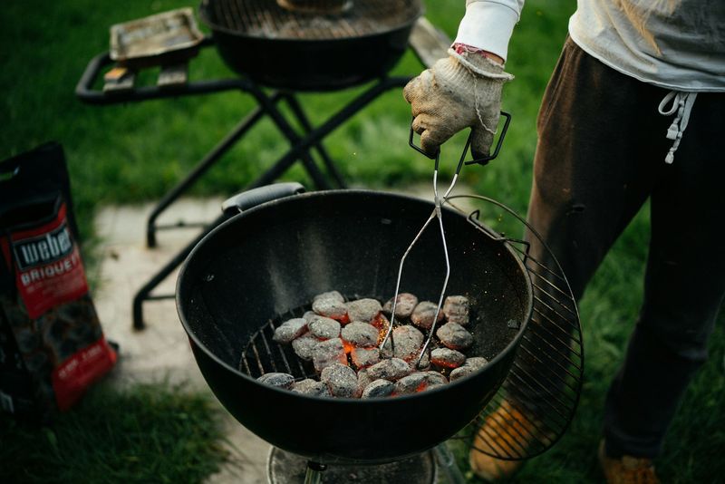 Hansket hånd holder en tang over glovarme kullbriketter i en rund grill