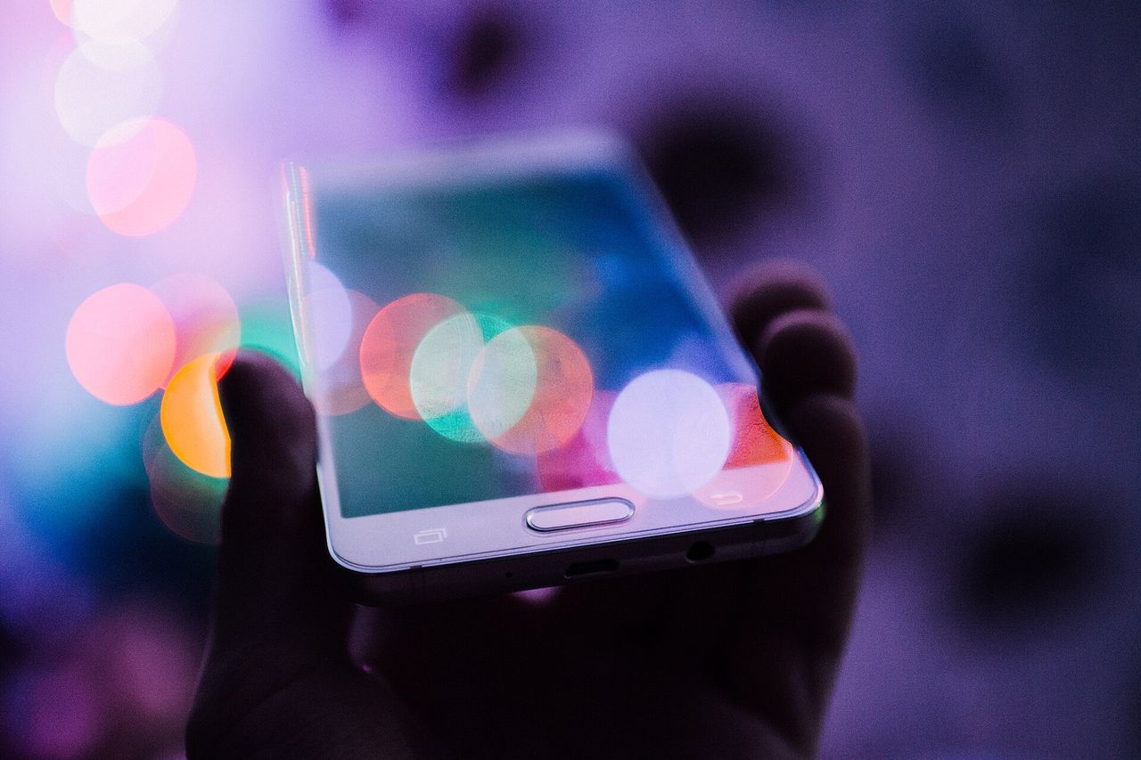 En hånd holder en smarttelefon foran fargerike, uskarpe lys