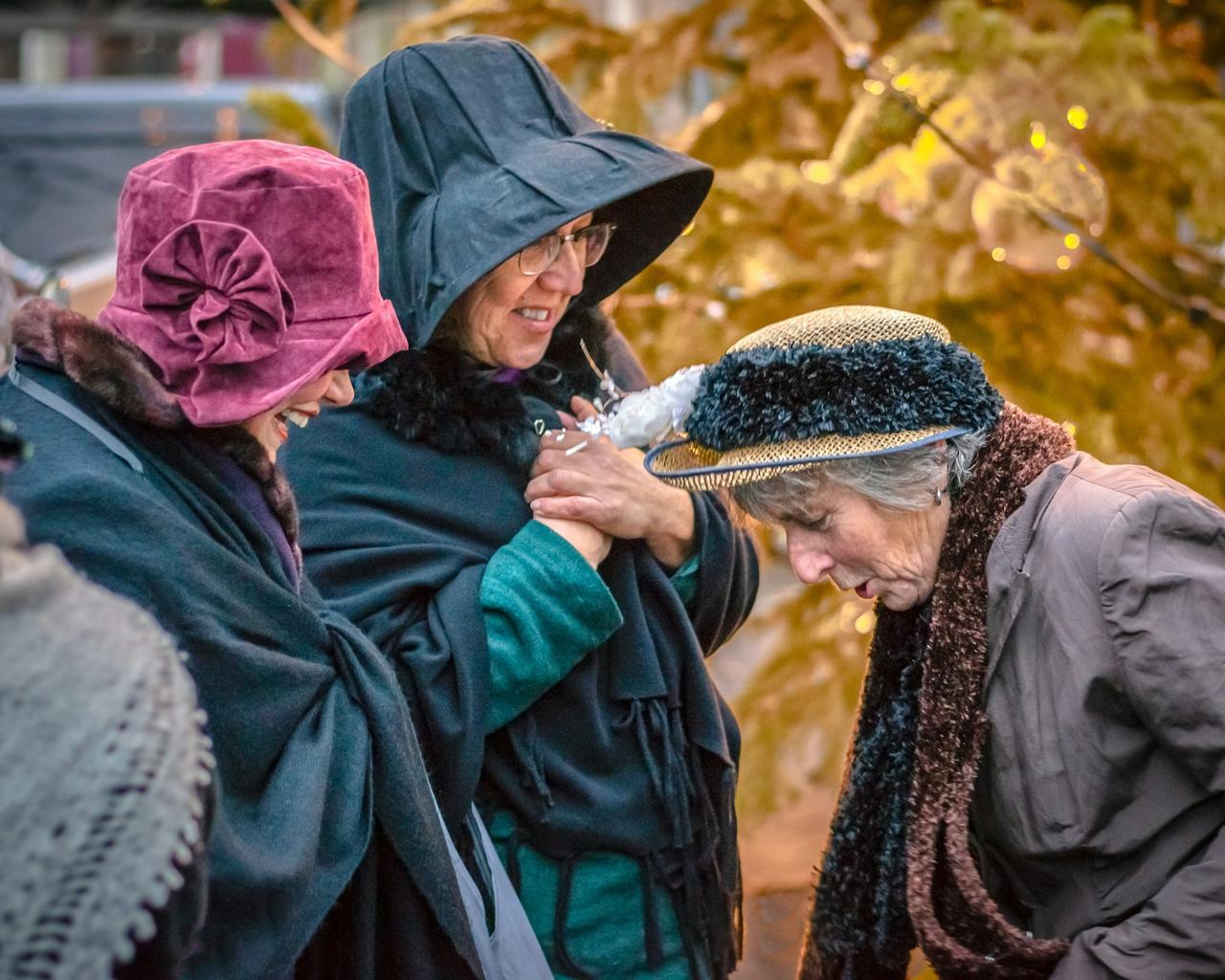 Tre eldre kvinner i hatter står tett sammen og ler i varmt høstlys